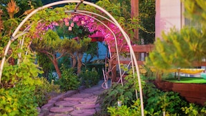 Ein Gartenweg mit einem hölzernen Bogen und einer Pergola, die mit rosa Blüten bedeckt ist.
