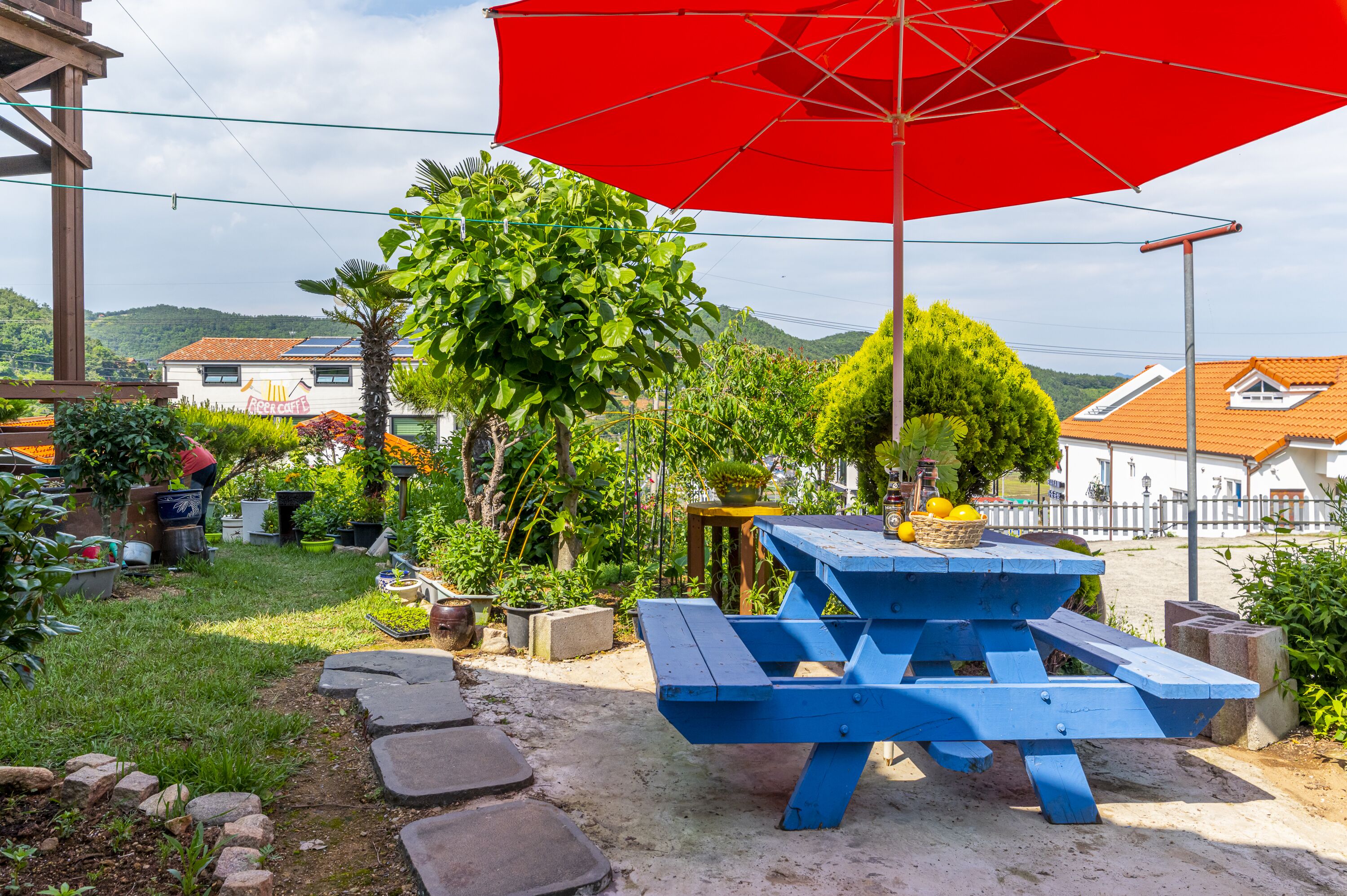 A blue picnic table with a red umbrella, surrounded by greenery and a white building with a red roof in the background.