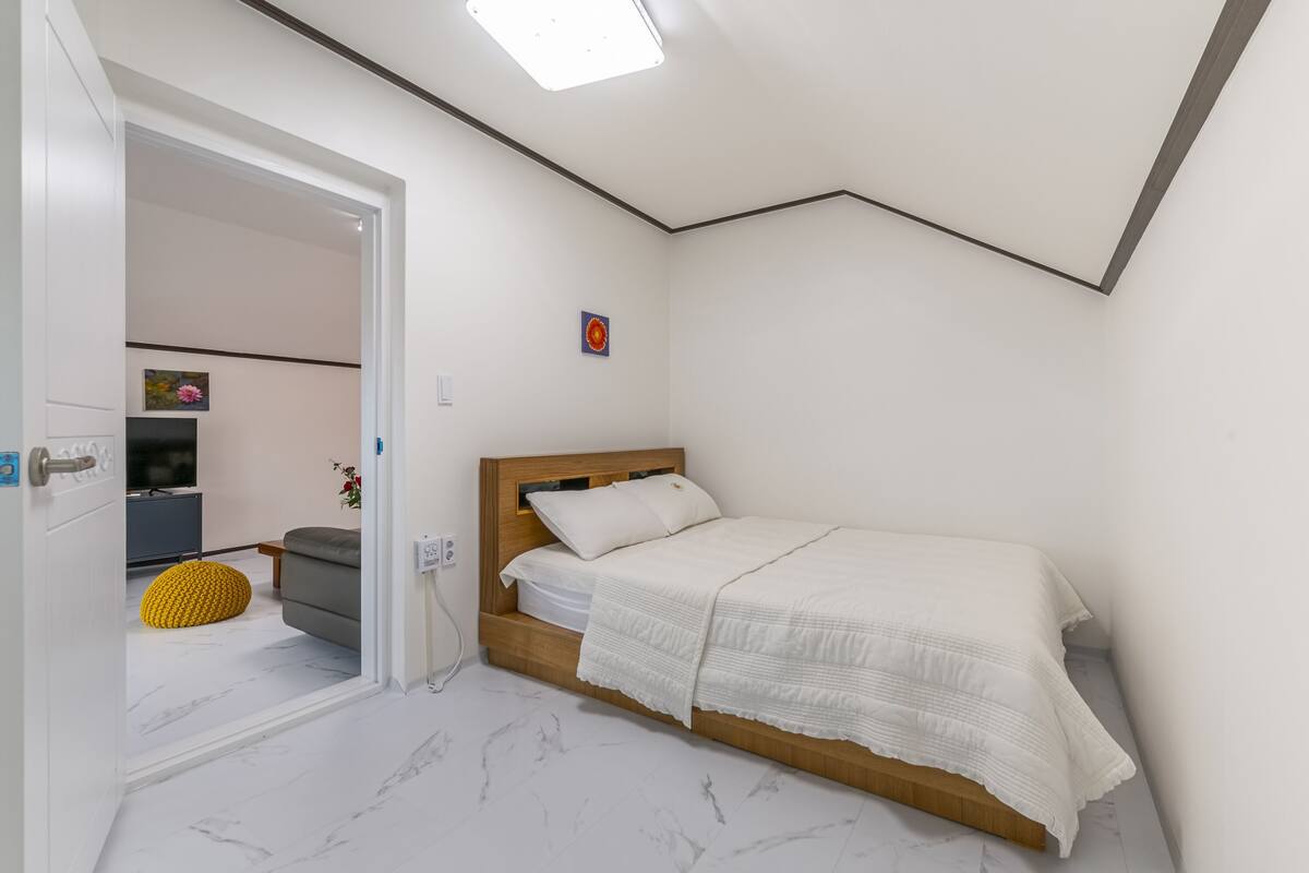 Un dormitorio con cama, cabecera de madera, un cuadro pequeño y un otoman amarillo.