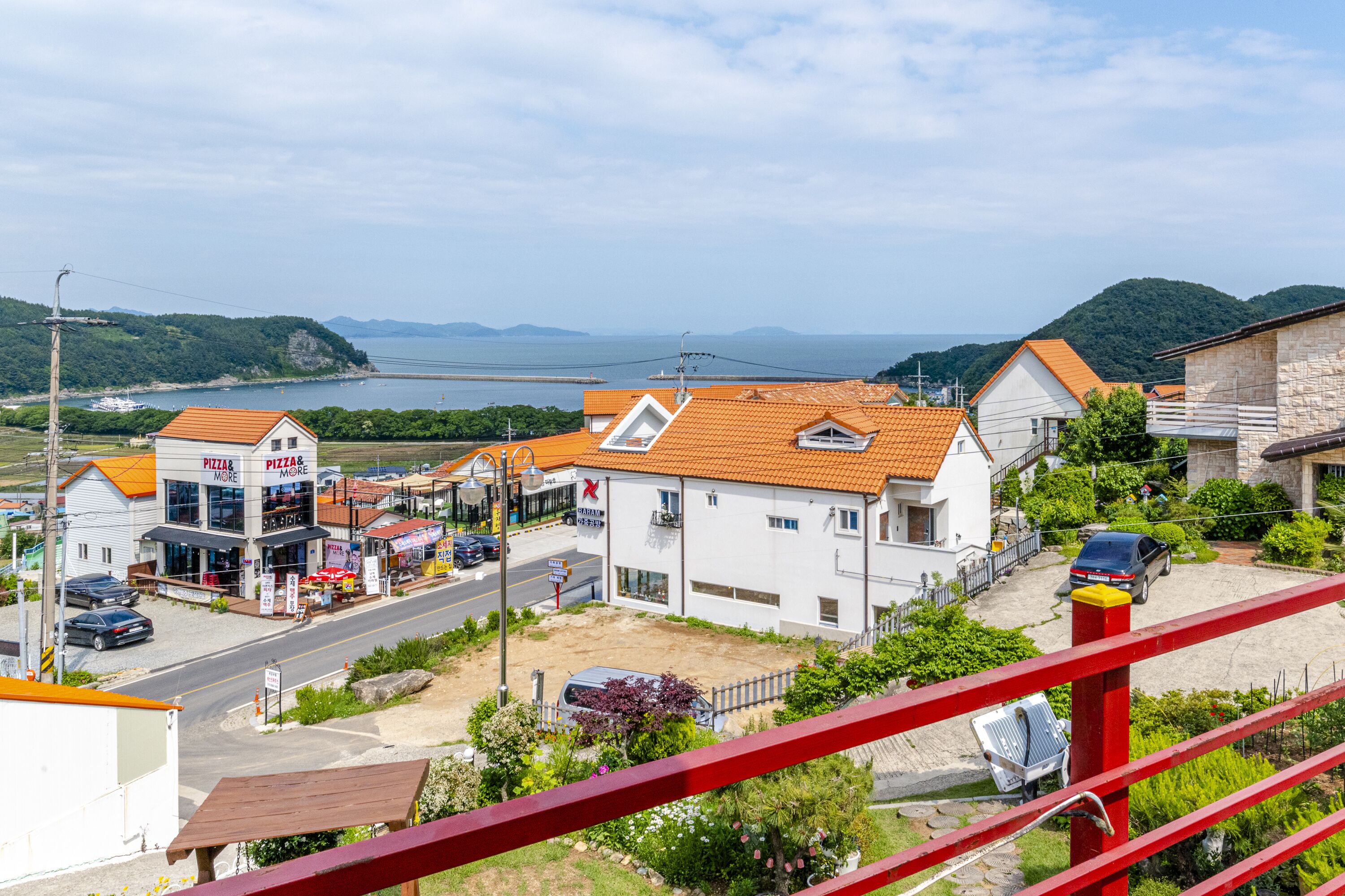 Uma vila costeira com um restaurante de pizza, um edifício branco com telhado vermelho e vista para o mar e colinas.