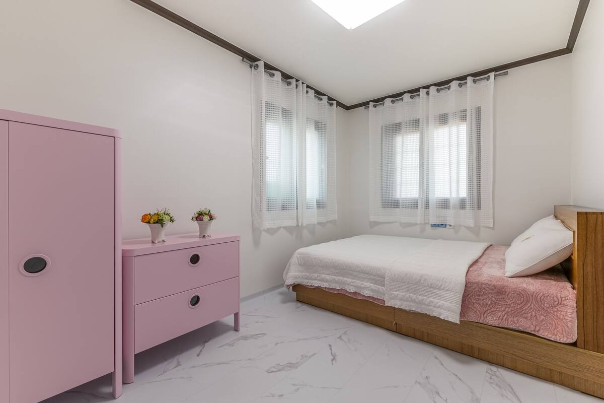 Un dormitorio con una cama, un tocador rosa con cajones, un cabecero de madera y una ventana con cortinas traslúcidas.