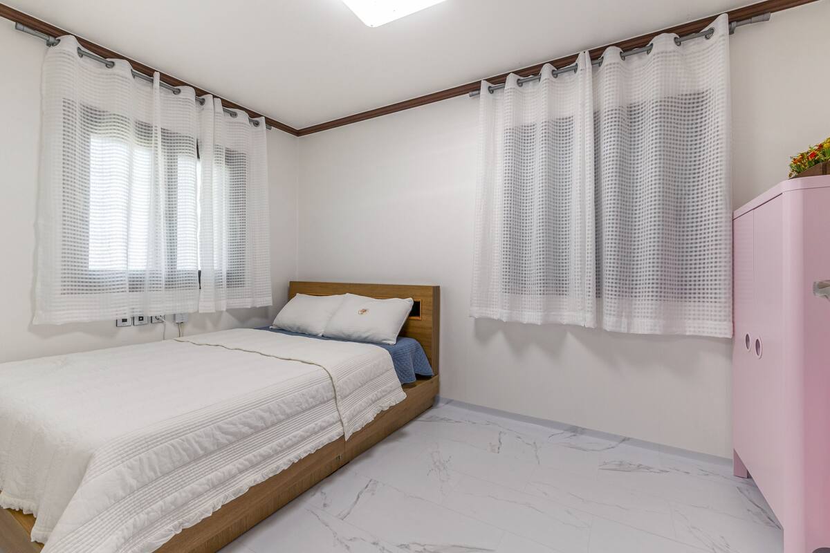 Un dormitorio con cama, cabecera de madera, un armario rosa y ventana con cortina blanca.