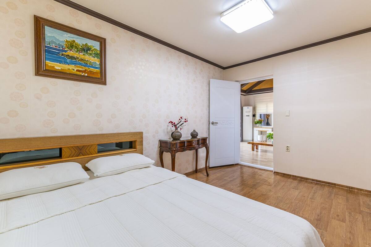 Un dormitorio con cama, una mesita de noche con un florero, un cuadro en la pared y una puerta que da a otra habitación.