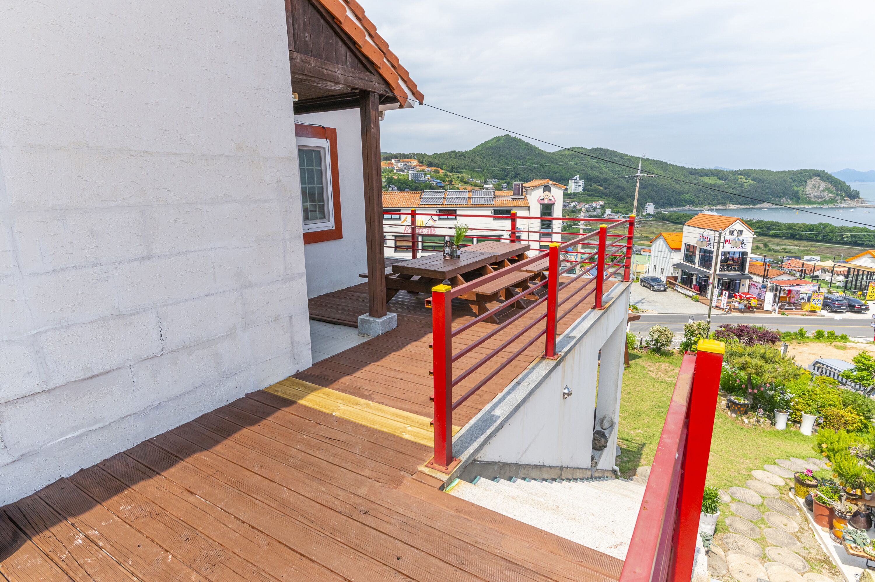 A wooden deck with a red railing overlooking a town with buildings and a view of the sea.