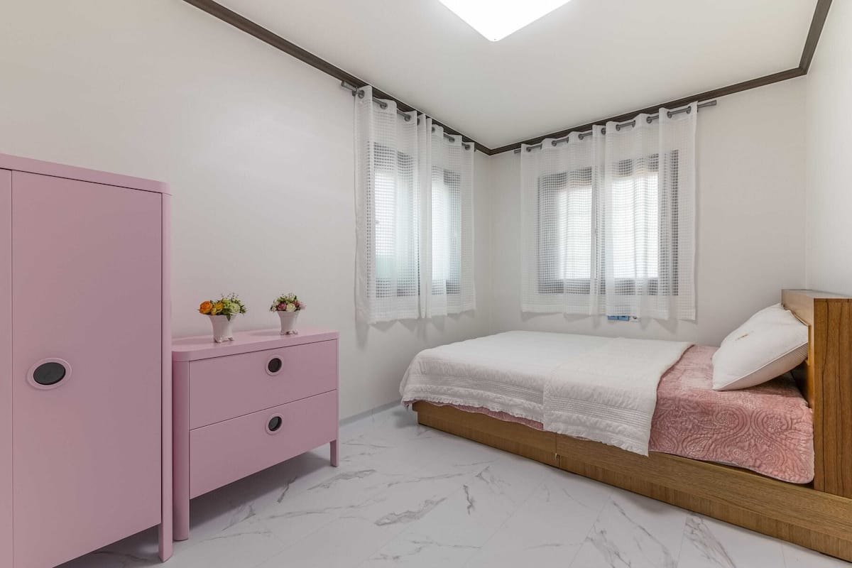 Un dormitorio con una cama, un tocador rosa y dos ventanas con cortinas traslúcidas.