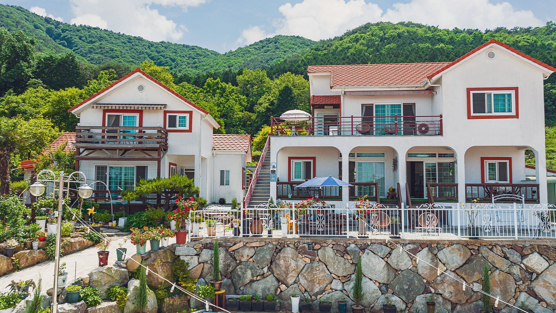 A two-story building with balconies, a stone wall in front, and a mountainous backdrop.