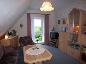 Ein gemütliches Wohnzimmer mit einer Couch, einem Couchtisch mit Spitzenplätzchen, einem Fernseher auf einem Schrank und einer Kommode mit Büchern und Dekoration.