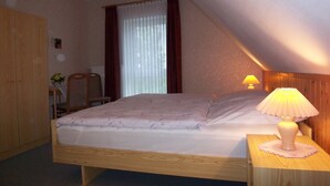 A bedroom with a wooden bed, a nightstand with a lamp, a chair, a wooden wardrobe, and a window with curtains.