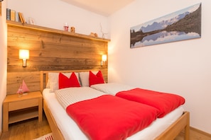 A double bed with red and white bedding, a wooden headboard, bedside lamps, a nightstand, and a framed picture of a mountain landscape.