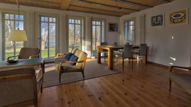 Un salon spacieux doté de planchers en bois, d’un canapé, d’une table à manger avec des chaises et de grandes fenêtres avec des rideaux.