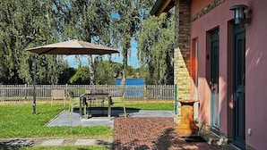 Un espace patio aménagé avec une table et des chaises, un grand parasol, et un chemin en briques menant à un bâtiment à la façade rose et doté d’un pilier en pierre.