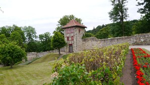A stone tower with a red-tiled roof, surrounded by a stone wall, a grassy area, and a flower bed with red flowers.