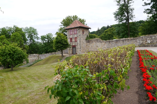 Una torre de pedra amb teulades vermelles, envoltada per una paret de pedra, una zona verda i un jardí amb flors vermelles.