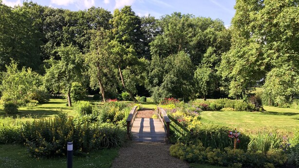Un sendero en un jardín que conduce a un puente de madera sobre un camino de grava, rodeado de abundante vegetación y plantas en flor.