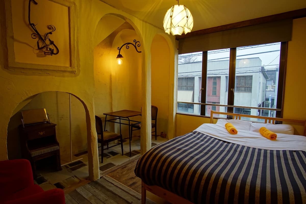 A bedroom with a bed, a desk, a chair, a piano, and a window with a view of buildings.
