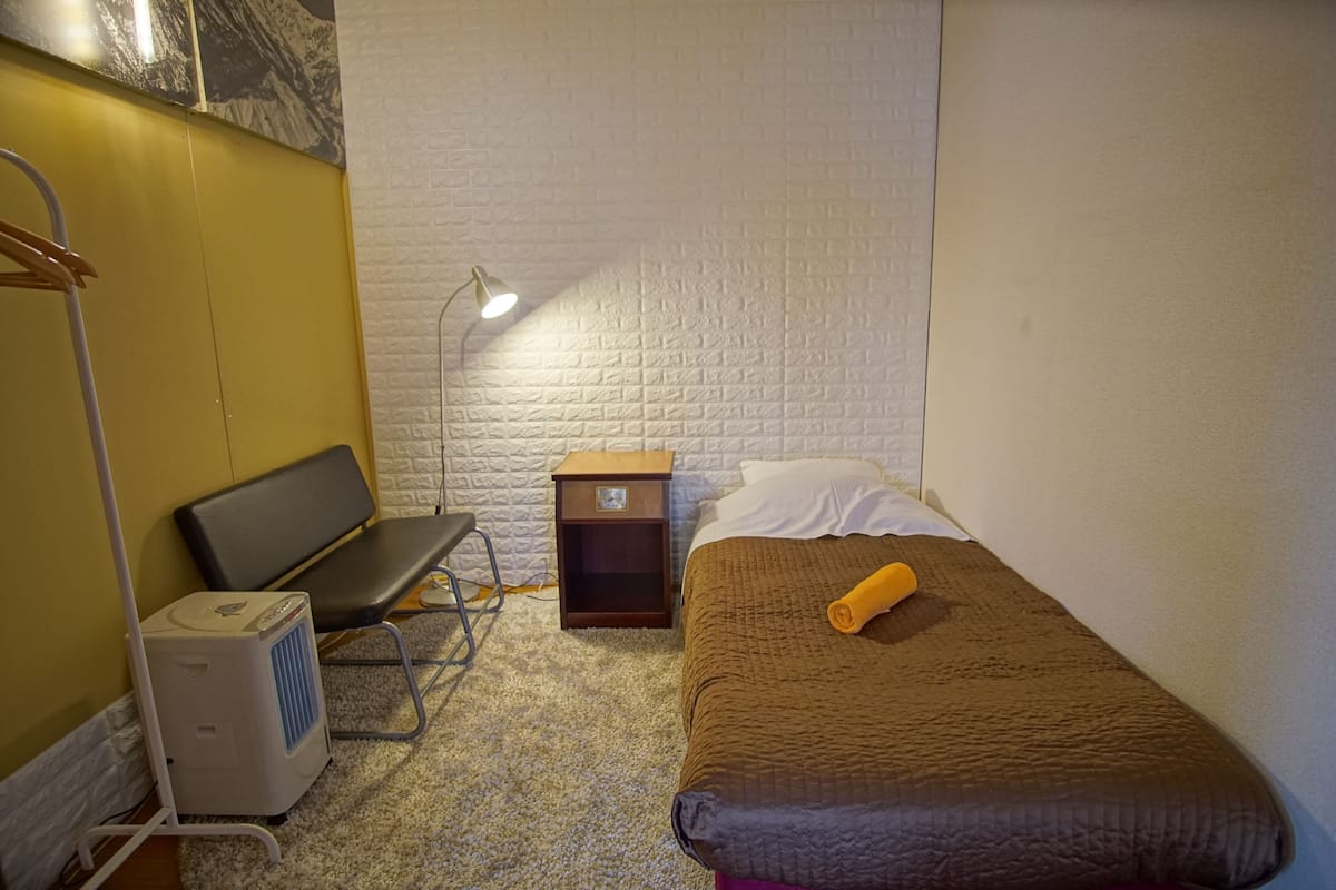 A small, neatly arranged bedroom with a bed, a bench, a desk, and a heater.