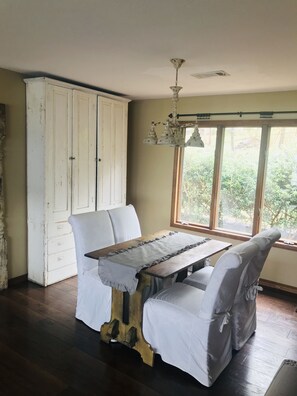 A dining area with a wooden table, white upholstered chairs, a white cabinet, and a view of greenery through large windows. - Beautiful Ozarks retreat on Bull Shoals Lake (Lakeview)