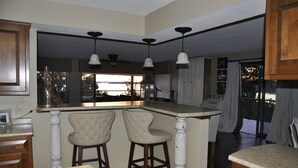 A kitchen with a bar area, two bar stools, pendant lights, and a view through a window.