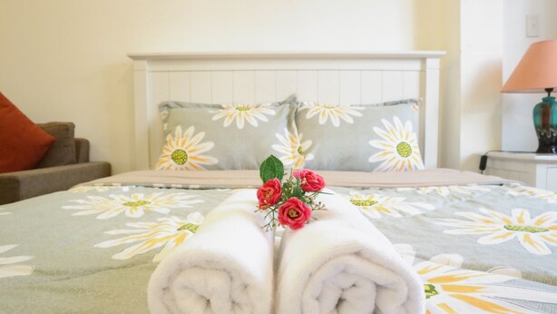 A neatly made bed with a floral bedspread, a pair of folded white towels, and a small bouquet of red flowers. - Smiley Apartment 3 (Ho Chi Minh City)