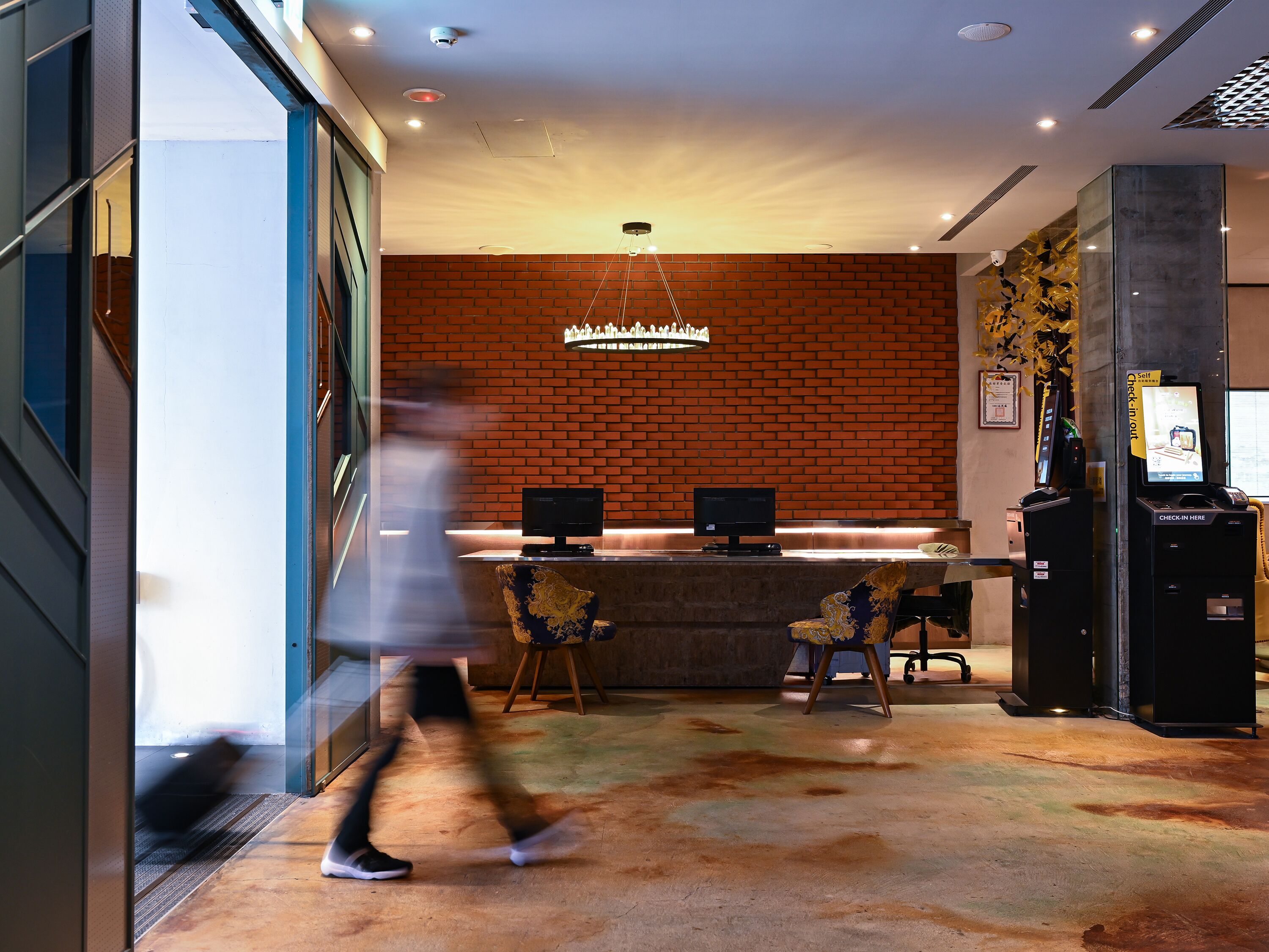 A modern hotel lobby with a brick wall, a reception desk, a chandelier, and a self-check-in kiosk.