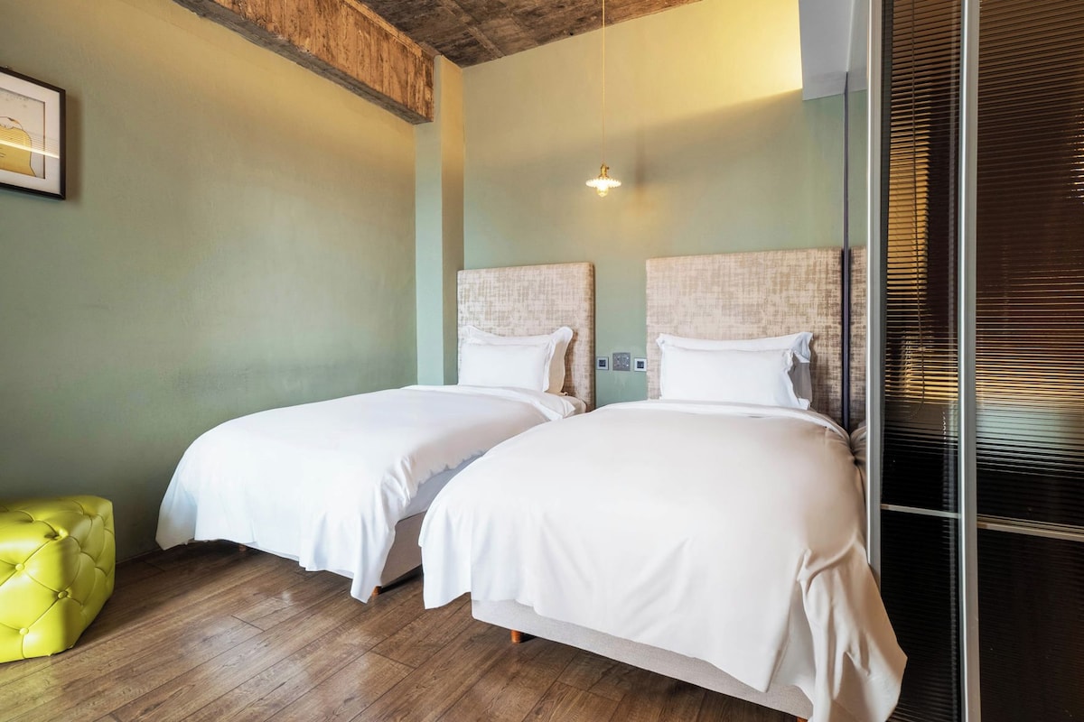 A hotel room with two beds, a yellow ottoman, and a wooden ceiling.
