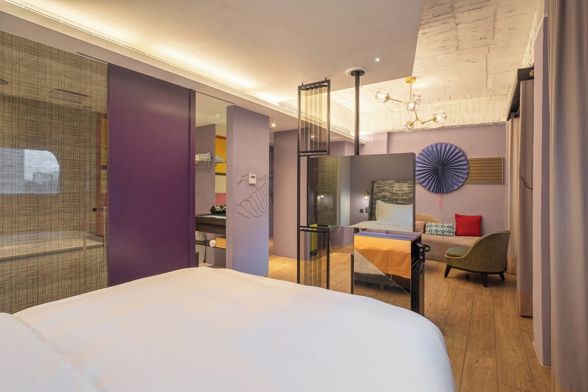 A modern hotel room with a large bed, a desk, a chair, and a dining area.