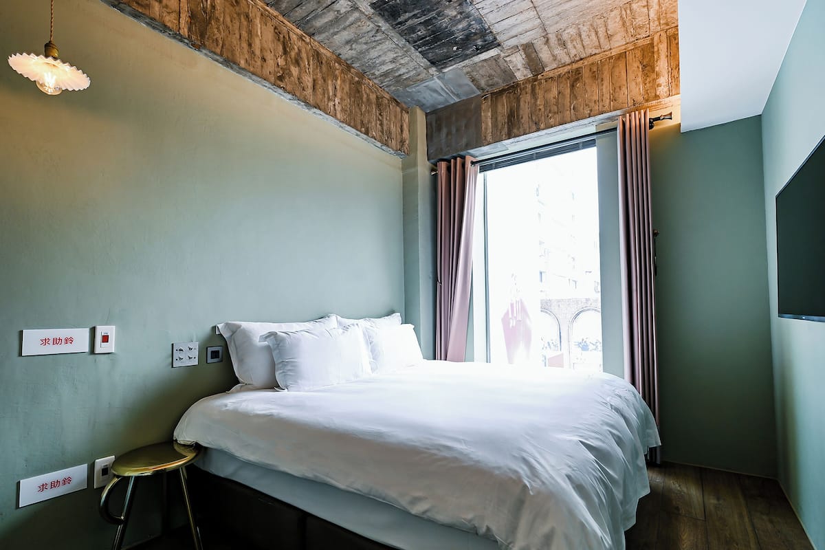 Una habitación de hotel con cama, un taburete, una lámpara y un ventanal con cortinas.