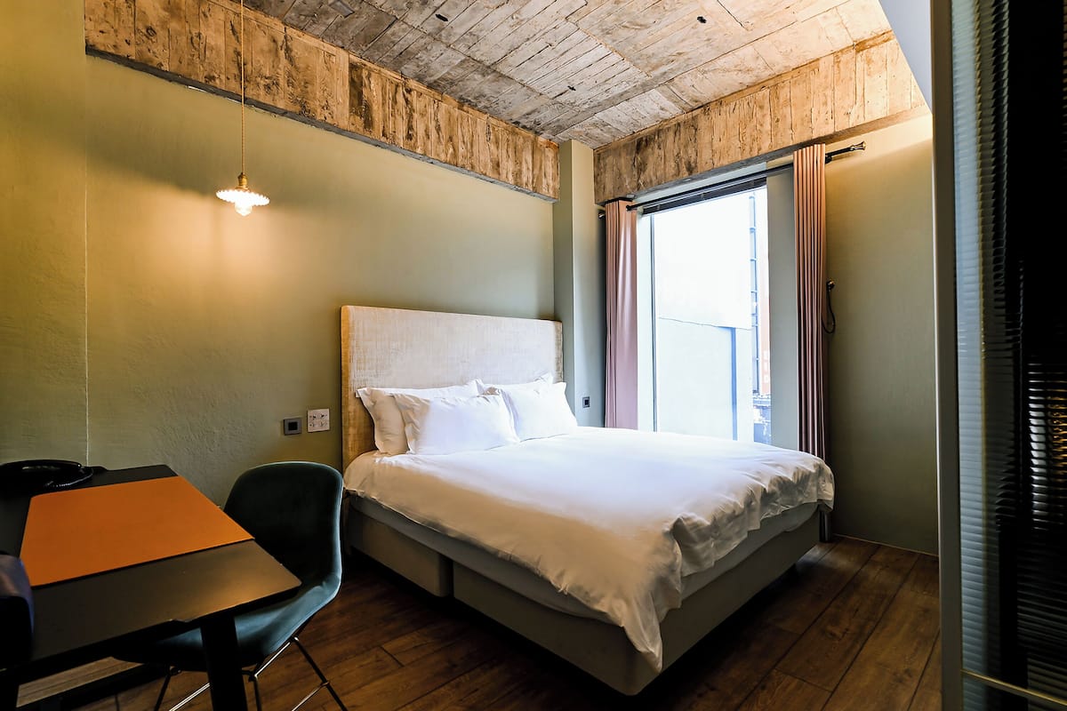Un dormitorio con techo de madera, una cama con ropa de cama blanca, un escritorio con silla y una ventana con cortinas.
