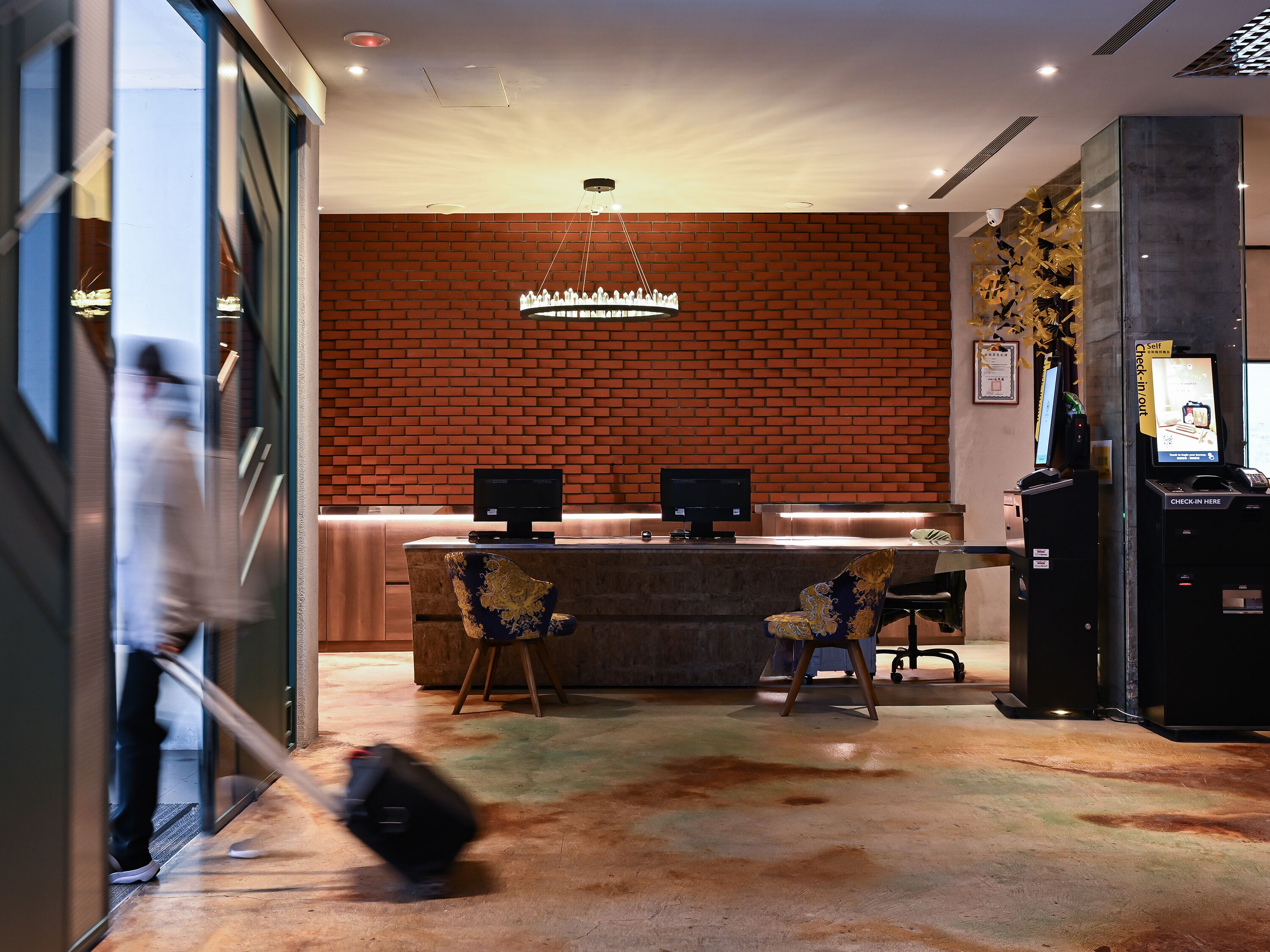 A hotel lobby with a brick wall, a reception desk, two monitors, a chandelier, and a self-check-in kiosk.