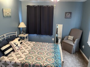 Un dormitorio con cama, una silla, una mesita de noche, una lámpara, un espejo y un cuadro de temática náutica además de un letrero.