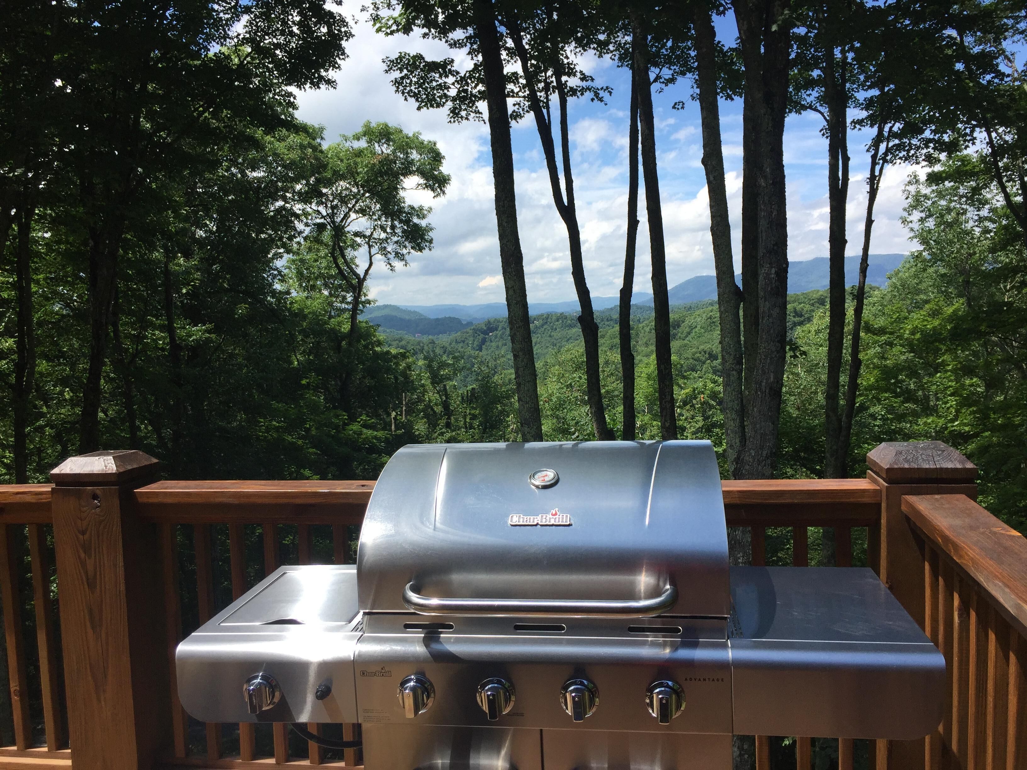 Una parrilla Char-Broil con cuatro quemadores, ubicada en una terraza de madera con vista a un bosque y montañas.
