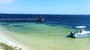 Um barco atracado em um cais que se estende por águas azul-claras, com uma praia de areia e uma cerca de madeira ao primeiro plano.