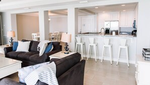 Una sala moderna con un sofá marrón oscuro, una mesa de centro blanca y una lámpara. Hay una zona de cocina con taburetes blancos y un refrigerador.