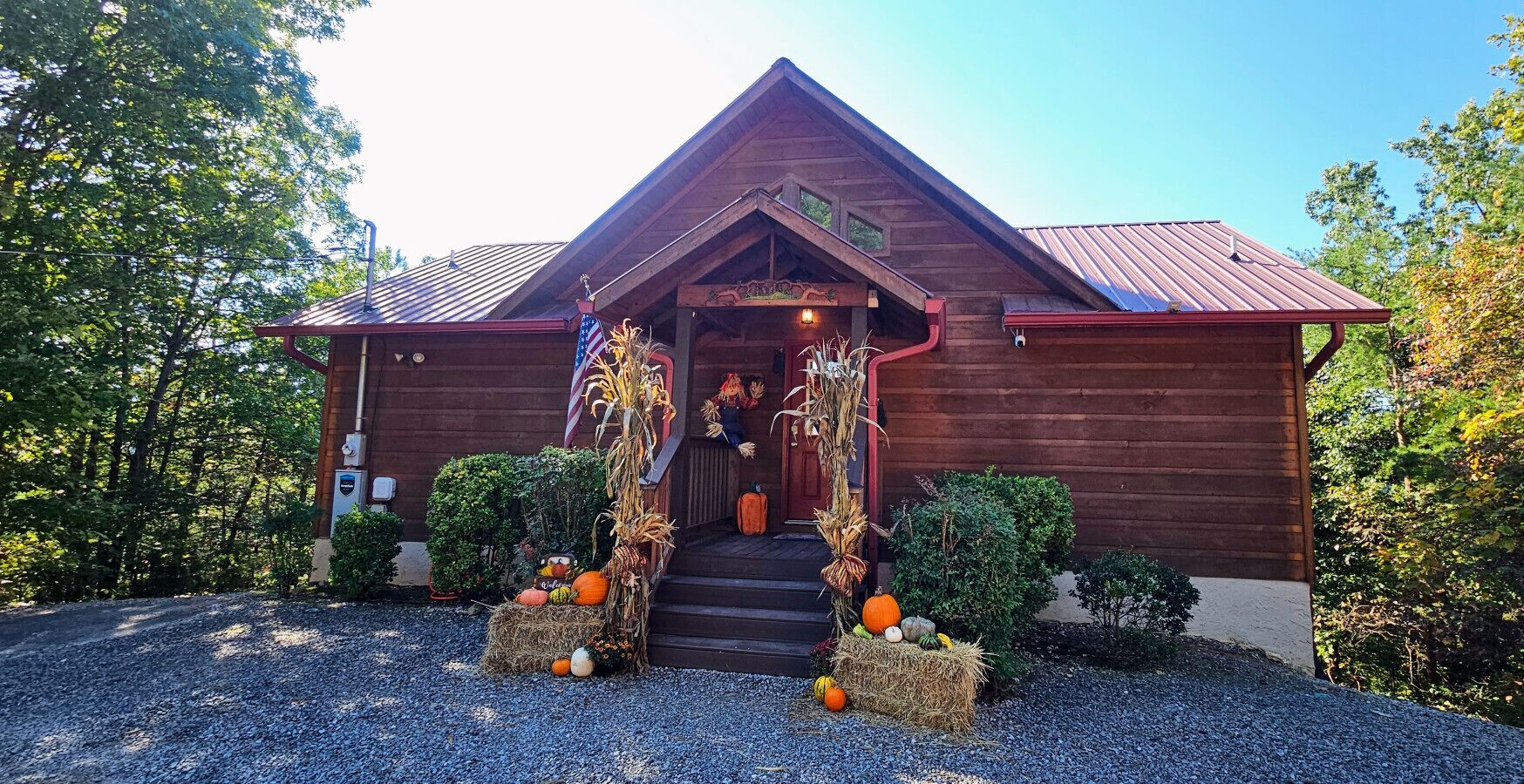 A rustic wooden cabin with a red roof, a front porch decorated with autumnal elements, and a gravel driveway.