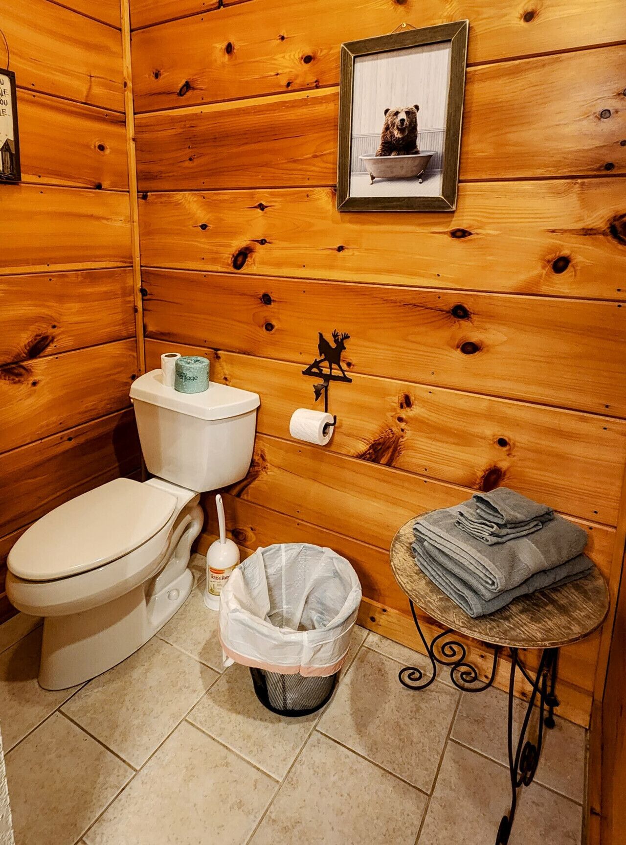 A wooden cabin bathroom with a toilet, a trash can, and a towel stack.