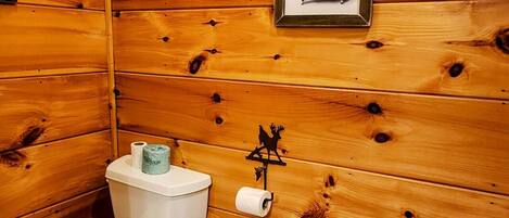 A wooden cabin bathroom with a toilet, a trash can, and a towel stack.