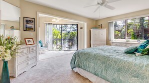 A bedroom with a large bed, a dresser, a chair, a window with a view of palm trees, and a balcony with a railing. - Spacious, Ground Floor, Waterfront Condo, Kanaloa at Kona Resort, Central Air (Kailua-Kona)