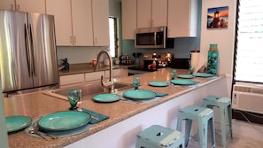A modern kitchen with stainless steel appliances, a granite countertop, and teal-colored plates and glasses set for dining. - Maui Tranquility ... Relax, Unwind and Feel Welcomed! (Kihei)