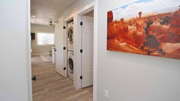 Una sala de lavandería con lavadora y secadora, un pasillo con una puerta que da a otra habitación, y un cuadro con paisaje de cañón en la pared.