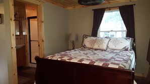 A bedroom with a wooden bed, a nightstand, a lamp, and a window with curtains. - Everything new in this beautiful secluded home. Air conditioning. Wifi. Firepit. (Lexington)