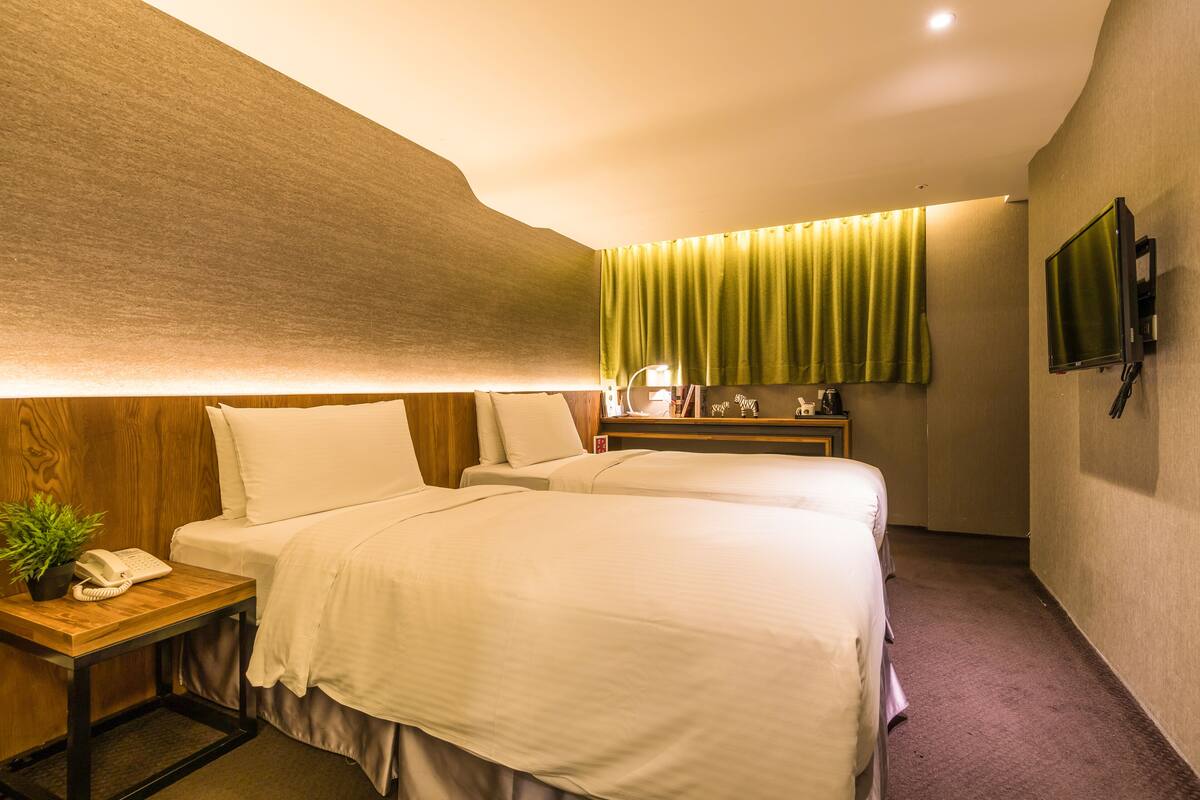 A hotel room with two beds, a bedside table with a telephone, a small plant, a nightstand with a lamp, and a television mounted on the wall.