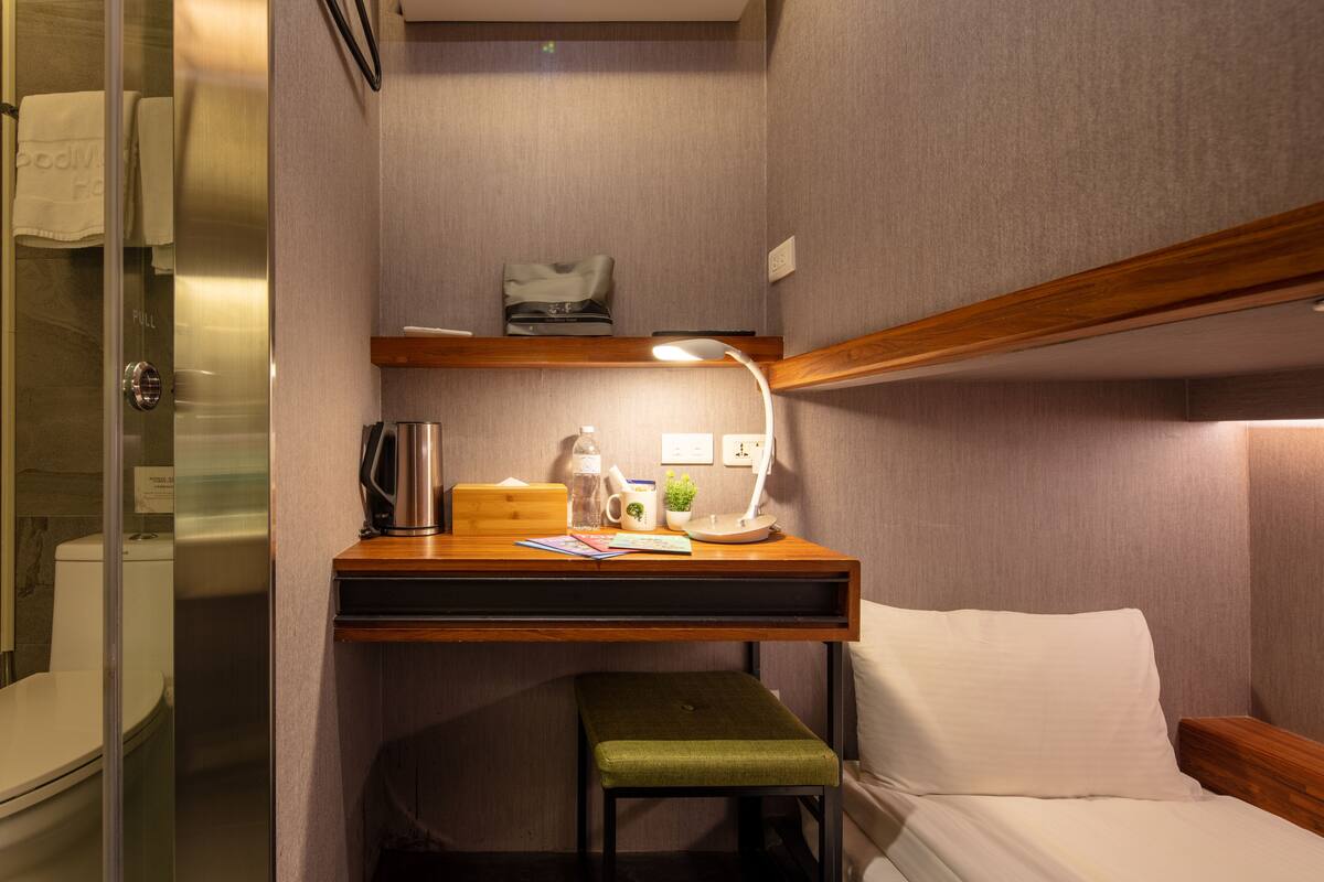 A compact hotel room with a bed, a small desk with a lamp, a green stool, and a shower area.