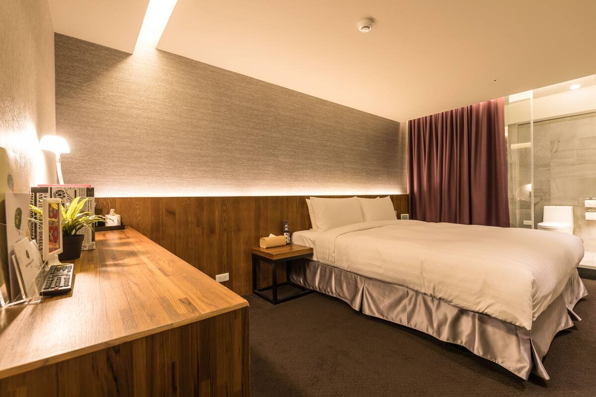 A hotel room with a wooden desk, a bed with white bedding, a small bedside table with a lamp, and a glass shower area.