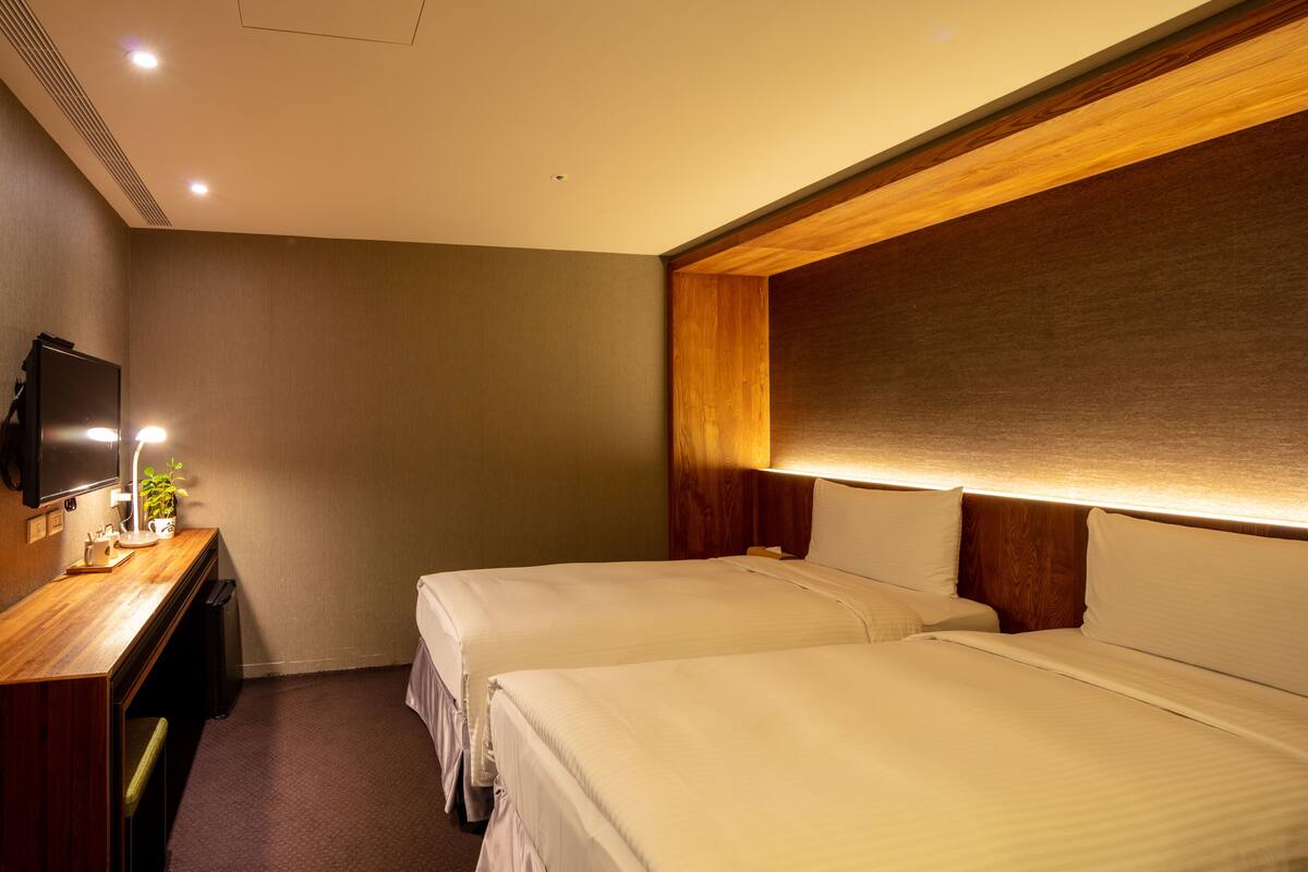 A hotel room with two beds, a desk with a lamp, and a television on the wall.
