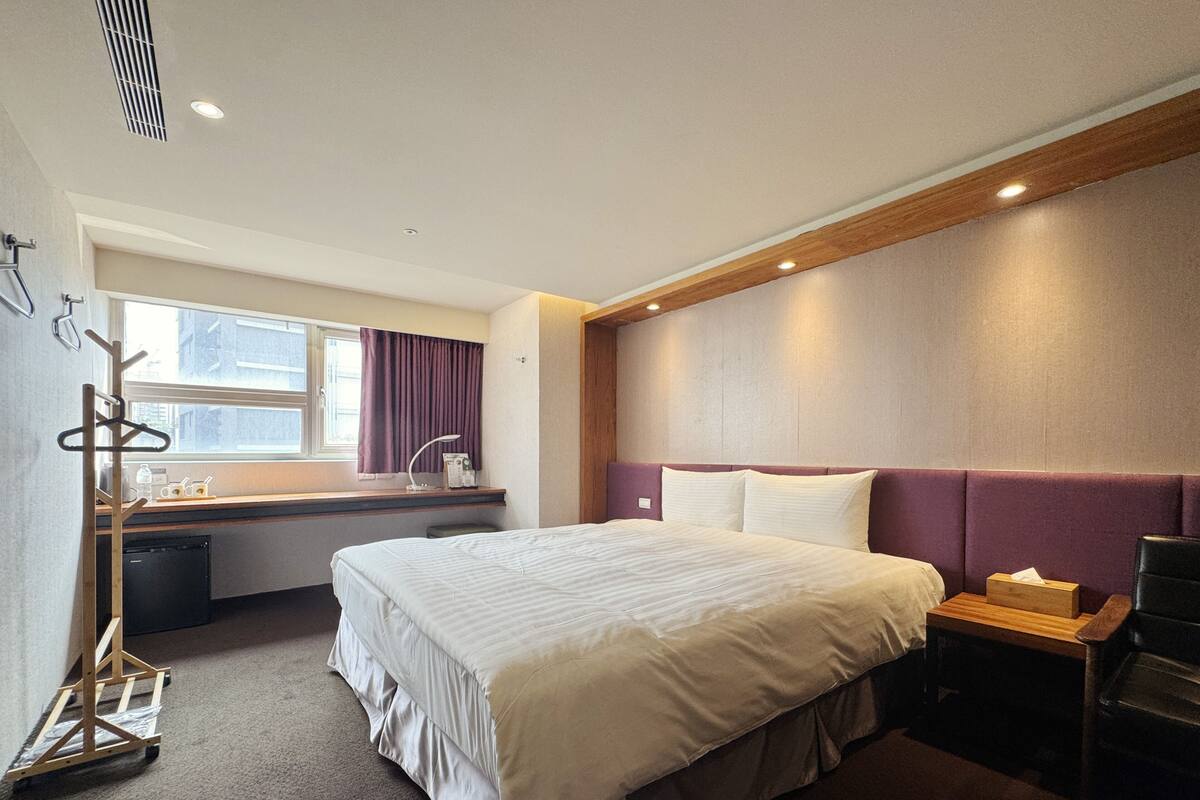 A hotel room with a bed, bedside table, chair, window, and a desk with a lamp.