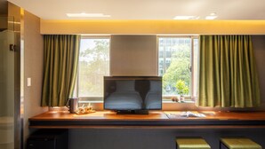 Habitación de hotel moderna con una gran televisión de pantalla plana sobre un escritorio de madera, dos bancos tapizados de color verde, una mesita con cafetera y una ventana con cortinas.