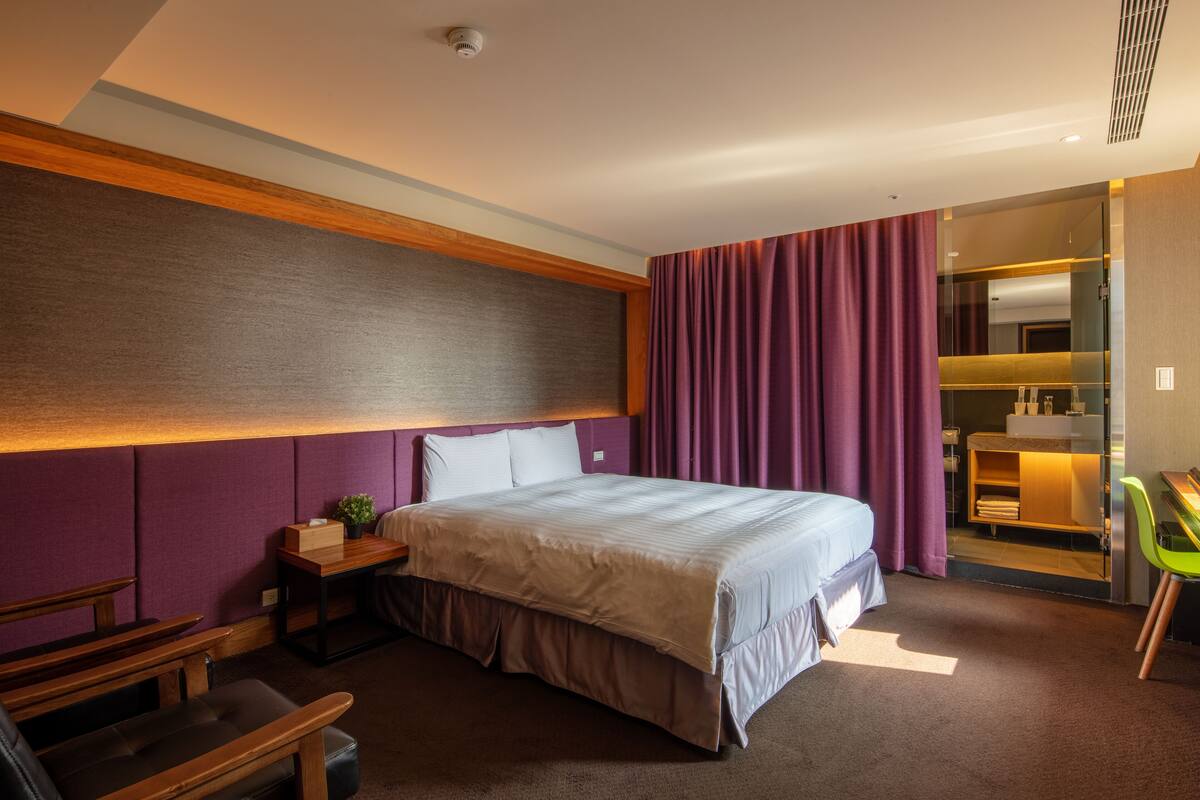 A modern hotel room with a large bed, a desk, a chair, and a small table.