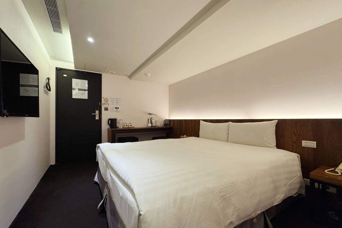 A hotel room with a bed, bedside table, lamp, and a wall-mounted phone.