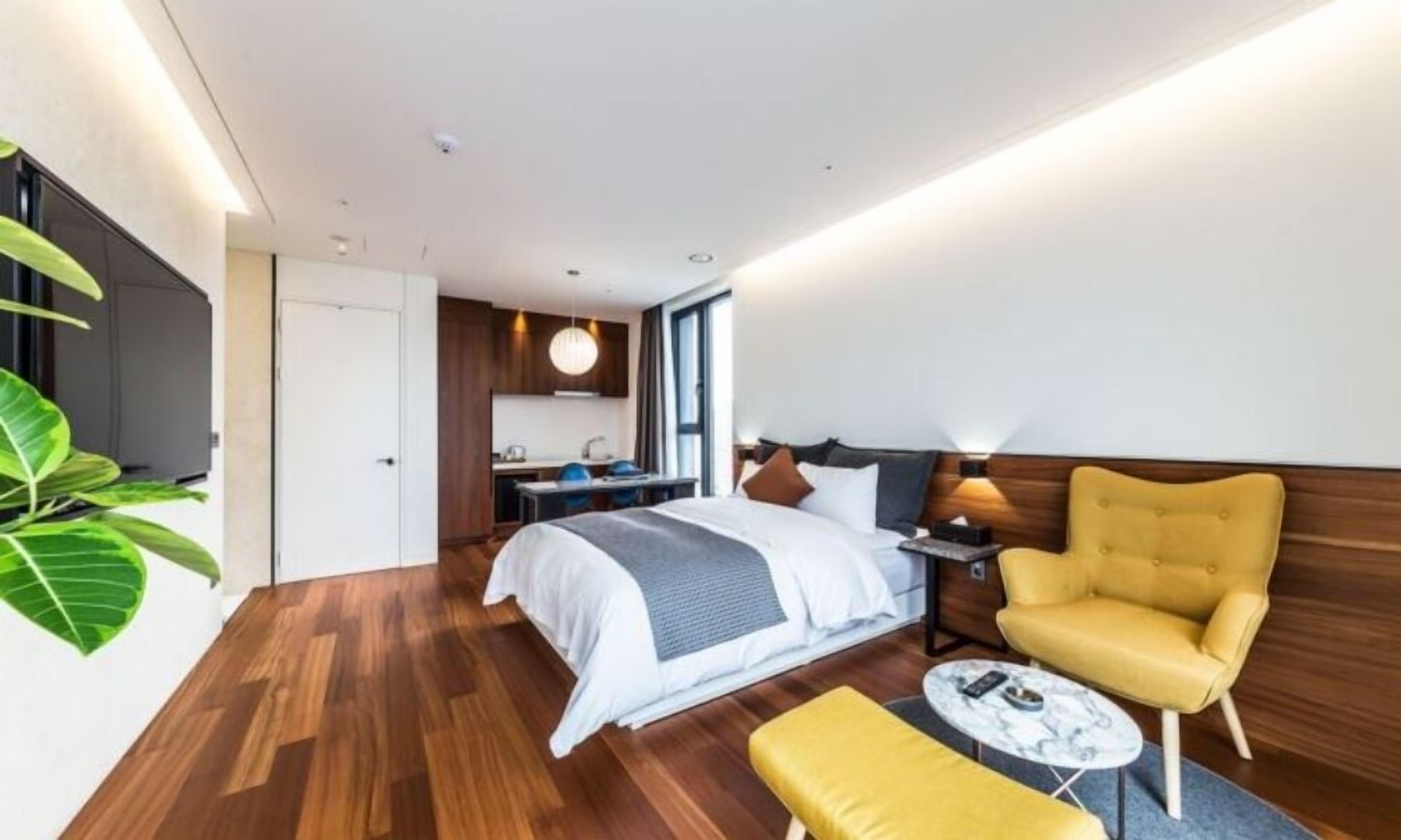 A modern hotel room with a wooden floor, a bed with white bedding, a yellow armchair, a small round table, and a view of the kitchen and bathroom through the door.
