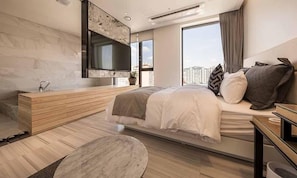 Une chambre moderne avec un grand lit, un téléviseur intégré, une coiffeuse en bois et une vue sur la ville.